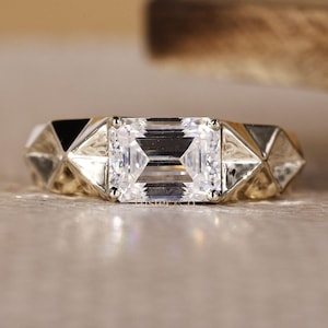 May include: A close-up of a gold ring with a large, rectangular, clear gemstone as the centerpiece. The band features geometric, triangular designs on either side of the gemstone, creating a modern and elegant look.