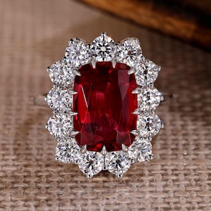 Puede incluir: Anillo de plata con una gran gema rectangular de color rojo intenso, rodeada por un halo de gemas más pequeñas, redondas y claras. El anillo se encuentra sobre un fondo neutro y texturizado.