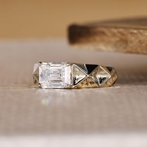 May include: A close-up of a geometric engagement ring with a large, rectangular, clear gemstone. The ring has a gold band with a silver geometric design on the sides. The ring is set against a neutral background.