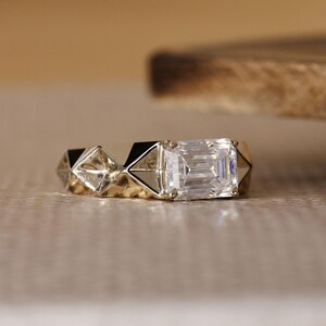 May include: A close-up of a gold ring with a large, rectangular, clear gemstone. The ring has a geometric design with triangular shapes along the band. The gemstone is the focal point, reflecting light.