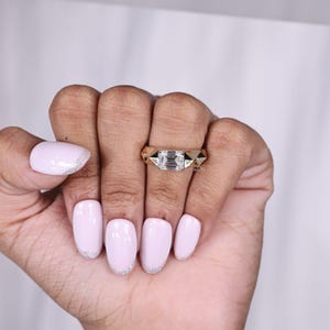 May include: A gold ring with a geometric design featuring three rectangular, clear stones. The ring has a unique shape with triangular accents on each side of the center stones. The ring is on a hand with light pink nail polish.