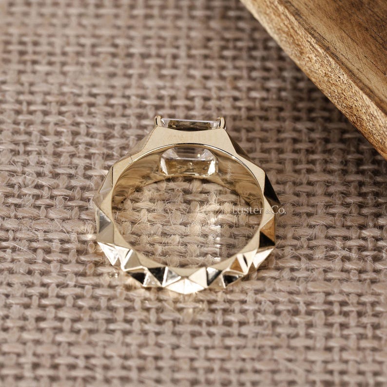 May include: A gold-colored ring with a geometric design. The ring features a square-cut, clear gemstone set in a unique, angular setting. The band has a faceted pattern, adding to its modern aesthetic.