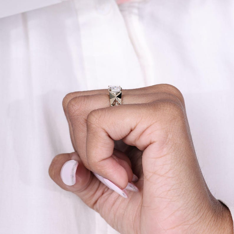 May include: A gold ring with a large, square, clear gemstone. The ring has a geometric design on the band. The ring is on a person's finger, with a white shirt in the background.