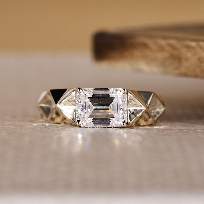 May include: A close-up of a gold ring with a large, rectangular, clear gemstone as the centerpiece. The band features geometric, triangular designs on either side of the gemstone, creating a modern and elegant look.