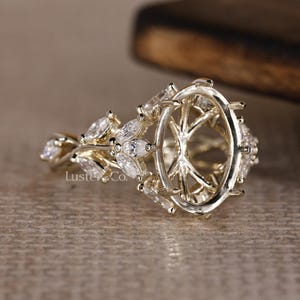 May include: A gold-toned ring setting with an oval center and intricate floral-inspired accents. The ring features multiple small, clear stones and the text "Luster & Co."