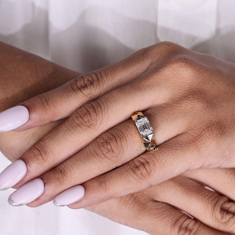 May include: A gold ring with a rectangular, clear gemstone set in the center. The ring has a geometric design with triangular accents. The ring is on a person's finger, with a white background.