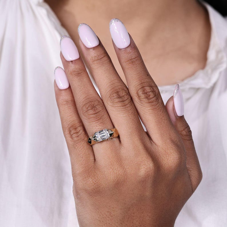 May include: A gold ring with a rectangular, emerald-cut center stone. The ring has black accents on the sides and is displayed on a hand with light pink nail polish.