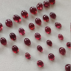 Może przedstawiać: Kolekcja owalnych, czerwonych kamieni szlachetnych. Kamienie szlachetne są rozrzucone na białym tle.
