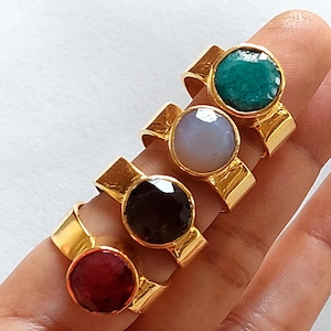 Peut inclure: Quatre bagues en or avec des pierres précieuses de différentes couleurs. Les bagues sont empilées les unes sur les autres. Les pierres précieuses sont rouge, noire, blanche et verte.