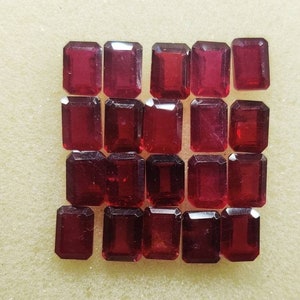 May include: A collection of 18 red gemstone rectangular cut cabochons. The gemstones are arranged in a grid pattern on a white surface.