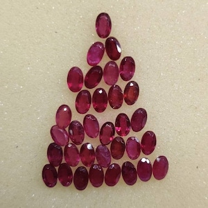 Könnte beinhalten: Eine Sammlung von 20 ovalen, tief roten Edelsteinen. Die Edelsteine sind in einer dreieckigen Form auf einer weißen Oberfläche angeordnet.