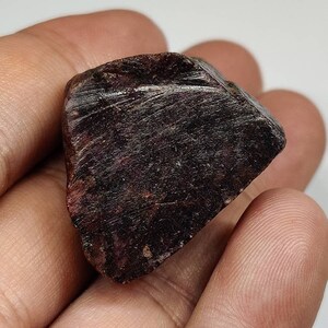 May include: A dark red ruby gemstone with a rough, natural surface. The ruby is a deep, rich red colour and has a slightly irregular shape.