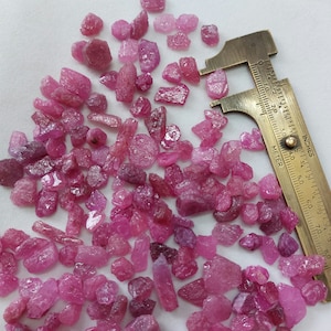 May include: A close-up of a collection of pink ruby gemstones of various sizes and shapes. The gemstones are spread out on a white surface and measured with a ruler in inches.