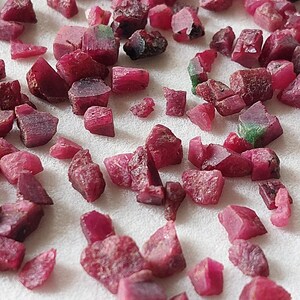 May include: A scattering of raw ruby gemstones in various sizes and shapes. The gemstones are a deep red colour, with some showing touches of green. The rough texture and natural forms of the rubies are visible, spread across a white surface.