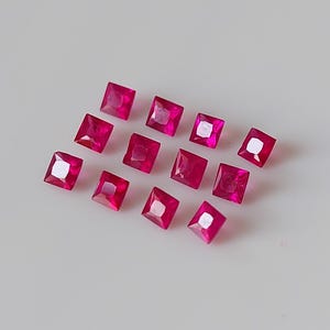 Peut inclure: Douze pierres précieuses de rubis de forme carrée, chacune d'une couleur rouge foncé, sont disposées en quinconce sur une surface blanche.