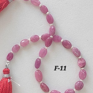 May include: A pink and white beaded necklace with a red tassel at each end. The necklace is made of oval-shaped beads and has a silver clasp. The text "F-11" is printed on a white background.