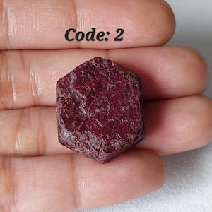 May include: A rough-cut, dark red ruby gemstone with a hexagonal shape. The surface of the ruby has a textured appearance. The text "Code: 2" is visible in the image.