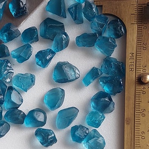 May include: A collection of teal blue gemstone roughs, some with sharp edges, scattered on a white surface. A ruler is visible in the background.