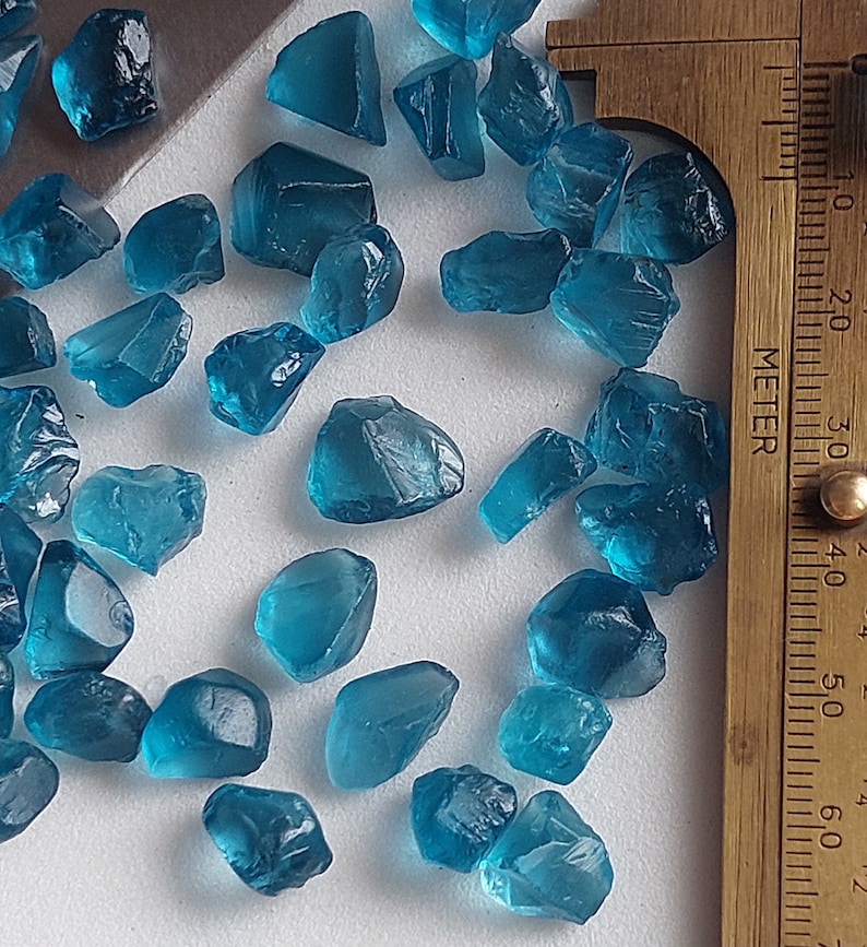 May include: A collection of teal blue gemstone roughs, some with sharp edges, scattered on a white surface. A ruler is visible in the background.