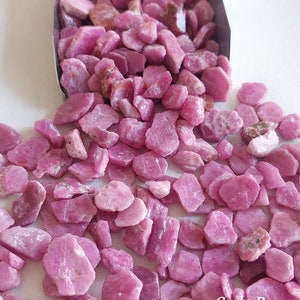 Könnte beinhalten: Nahaufnahme eines Haufens rosafarbener Rubin-Edelsteine. Die Rubine sind in verschiedenen Größen und Formen, und einige haben eine raue, unpolierte Textur. Die Rubine fallen aus einem Metallbehälter, und der Hintergrund ist weiß. Code B.