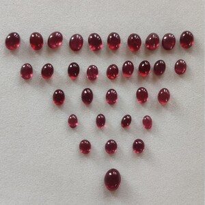 May include: A collection of 25 round, red gemstones arranged in a triangular pattern on a white surface.