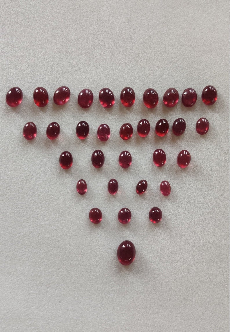 May include: A collection of 25 round, red gemstones arranged in a triangular pattern on a white surface.