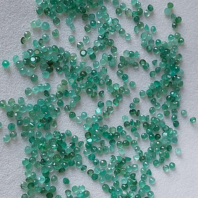 May include: A collection of small, faceted emerald gemstones in various shades of green. The round gemstones are polished, reflecting light. The gems are scattered across a neutral surface, suggesting a collection of loose stones.