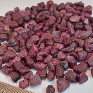 May include: A pile of small, rough, pink ruby stones. The stones are scattered on a white surface next to a ruler.