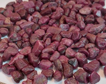 Peut inclure: Un tas de petites pierres de rubis roses brutes. Les pierres sont éparpillées sur une surface blanche à côté d'une règle.