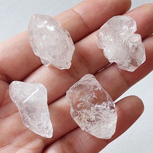 Peut inclure: Quatre amas de cristaux de quartz clairs de formes irrégulières. Les cristaux sont translucides et semblent être taillés à l'état brut, mettant en valeur les formations naturelles. Les cristaux sont tenus dans une main, sur fond neutre.