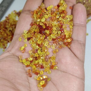 May include: A handful of small, polished gemstones in shades of yellow, orange, and red. The gems are irregularly shaped and appear to be scattered across a palm. The gemstones are likely used for jewelry making or collecting.