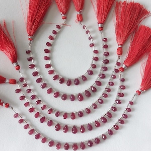 May include: Strands of deep red, teardrop-shaped gemstones strung on silver wire, with matching red tassels at the ends. The gemstones are arranged in curved lines, creating a layered effect. The background is plain white.