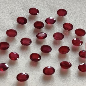 May include: A collection of oval-shaped, red gemstones on a white surface. The gemstones are arranged in a loose, scattered pattern.