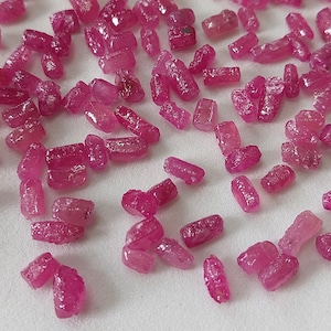 Op de afbeelding: Een verzameling kleine, ruwe, roze robijn edelstenen. De robijnen zijn van verschillende groottes en vormen, sommige zijn meer rechthoekig en andere meer rond. De robijnen zijn verspreid over een wit oppervlak.