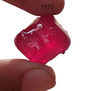 Puede incluir: Una piedra preciosa facetada de color rojo intenso con forma cuadrada. La piedra preciosa se sostiene en una mano, y el número 1971 es visible en el fondo.