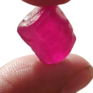 Könnte beinhalten: Ein leuchtend rosa Edelstein mit einer rauen, facettierten Oberfläche wird zwischen zwei Fingern gehalten. Der Edelstein ist etwa 2,5 cm groß. Die Nummer 1020 ist im Hintergrund sichtbar.