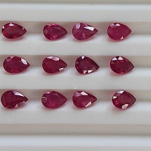 May include: Twelve pear-shaped ruby gemstones arranged in three rows of four on a white surface. The rubies are a deep red color and have a glossy finish.