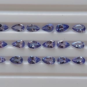 May include: A collection of 18 pear-shaped gemstones, arranged in three rows of six. The gemstones are a light purple color with a slight shimmer.