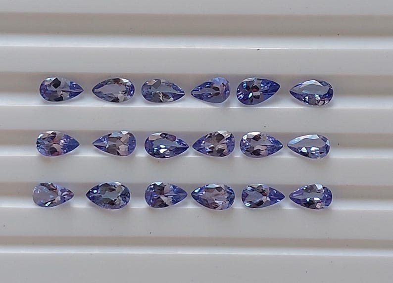 May include: A collection of 18 pear-shaped gemstones, arranged in three rows of six. The gemstones are a light purple color with a slight shimmer.