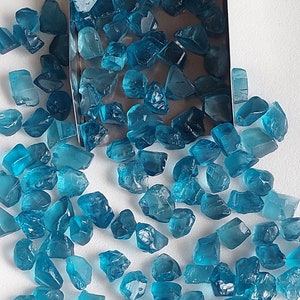 May include: A collection of loose, faceted, teal blue gemstones. The gemstones are scattered on a white surface and in a metal tray.