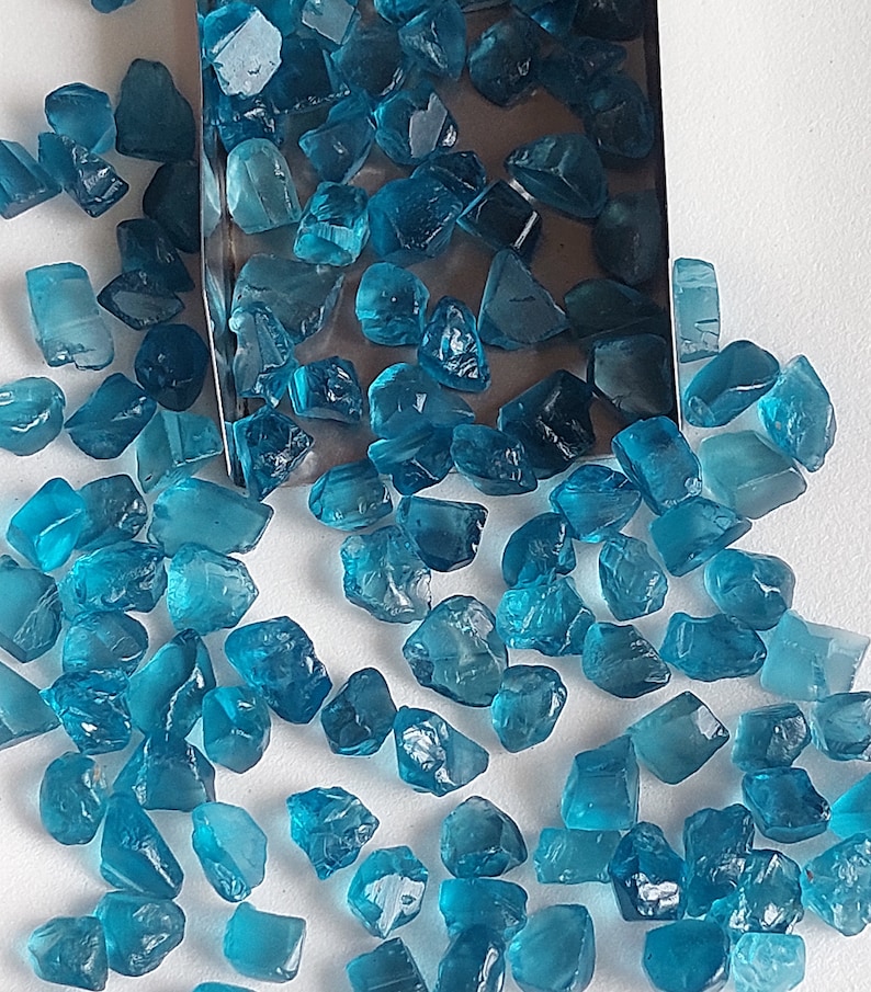 May include: A collection of loose, faceted, teal blue gemstones. The gemstones are scattered on a white surface and in a metal tray.