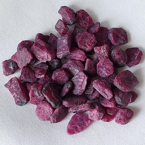 Pode incluir: Um monte de pedras preciosas de rubi em bruto. As pedras são de cor vermelha profunda com uma superfície ligeiramente texturizada. As pedras preciosas têm formas irregulares e tamanhos variados, algumas exibindo uma estrutura cristalina.