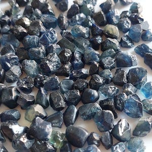 Può includere: Una collezione di zaffiri grezzi, non tagliati, in varie sfumature di blu e verde acqua. Le pietre preziose, dalle forme naturali e irregolari, sono sparse su una superficie bianca. Gli zaffiri variano in dimensioni e presentano una lucentezza sottile.