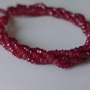 Puede incluir: Un hilo de cuentas de piedras preciosas de color rojo intenso y talladas, dispuestas en un bucle circular. Cada pequeña cuenta circular presenta múltiples superficies planas que captan la luz, creando un efecto brillante en el cordón carmesí.