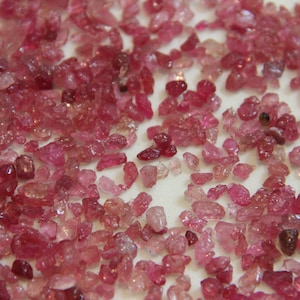 May include: A close-up view of a collection of pink ruby gemstones. The rough, uncut stones are scattered on a white surface.