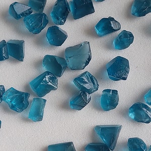May include: A collection of teal blue gemstone crystals of various shapes and sizes. The crystals are scattered on a white surface.
