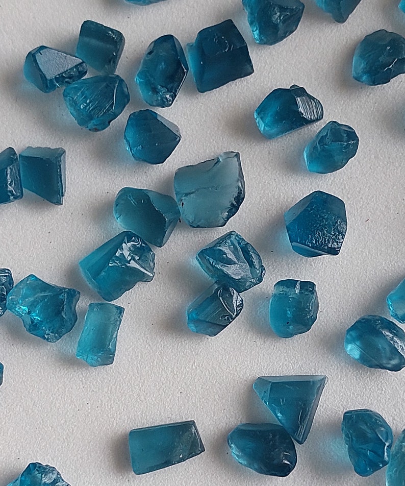 May include: A collection of teal blue gemstone crystals of various shapes and sizes. The crystals are scattered on a white surface.