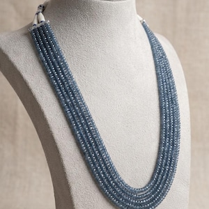 Puede incluir: Un collar de múltiples hilos que presenta cinco filas de pequeñas cuentas facetadas de color azul pizarra. Los hilos se reúnen en la parte posterior con un cierre de cordón blanco texturizado, creando un efecto de capas en cascada.