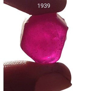 Puede incluir: Una piedra preciosa facetada de color rosa intenso con una superficie lisa y pulida. La piedra preciosa se sostiene entre dos dedos. El número 1939 es visible en el fondo.