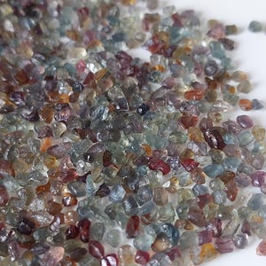 Puede incluir: Una colección de pequeños fragmentos de piedras preciosas en bruto y multicolores en tonos azul, rojo, morado y amarillo. Estas diminutas piedras translúcidas e irregulares tienen una textura cristalina facetada sobre un fondo blanco.